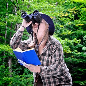 woman in the woods looking through binoculars and holding a book
